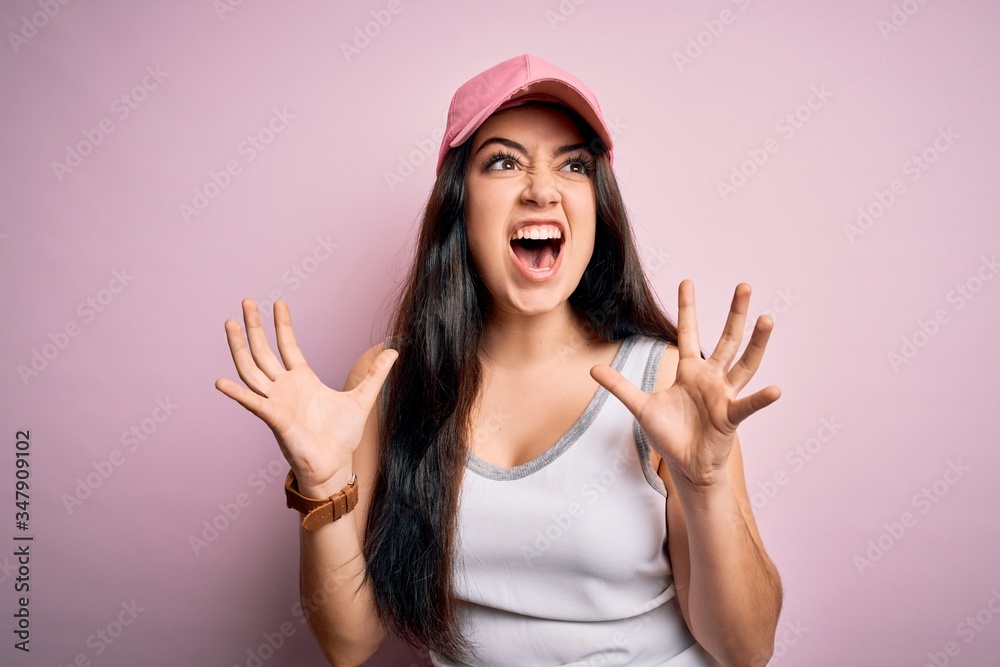Young brunette woman wearing casual sport cap over pink background crazy and mad shouting and yelling with aggressive expression and arms raised. Frustration concept. Young brunette woman wearing casual sport cap over pink background crazy and mad shouting and yelling with aggressive expression and arms raised. Frustration concept.