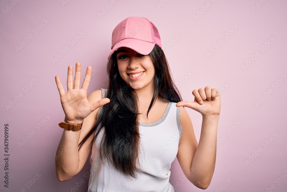 Young brunette woman wearing casual sport cap over pink background showing and pointing up with fingers number six while smiling confident and happy. Young brunette woman wearing casual sport cap over pink background showing and pointing up with fingers number six while smiling confident and happy.