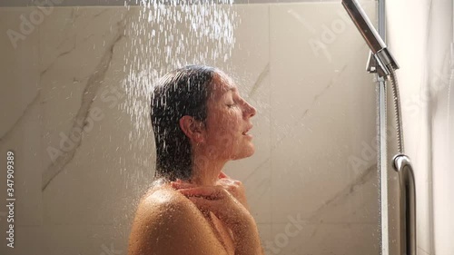 young woman stands under heavy water streams and washes face in home bathroom shower cabin slow motion close view