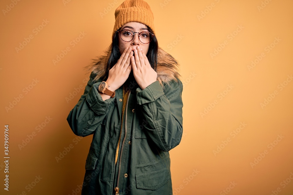 Young brunette woman wearing glasses and winter coat with hat over yellow isolated background shocked covering mouth with hands for mistake. Secret concept. Young brunette woman wearing glasses and winter coat with hat over yellow isolated background shocked covering mouth with hands for mistake. Secret concept.