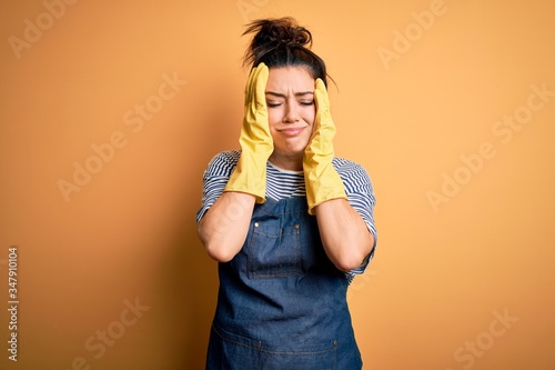 Fotografie Young brunette cleaner woman wearing housekeeping gloves over yellow background suffering from headache desperate and stressed because pain and migraine