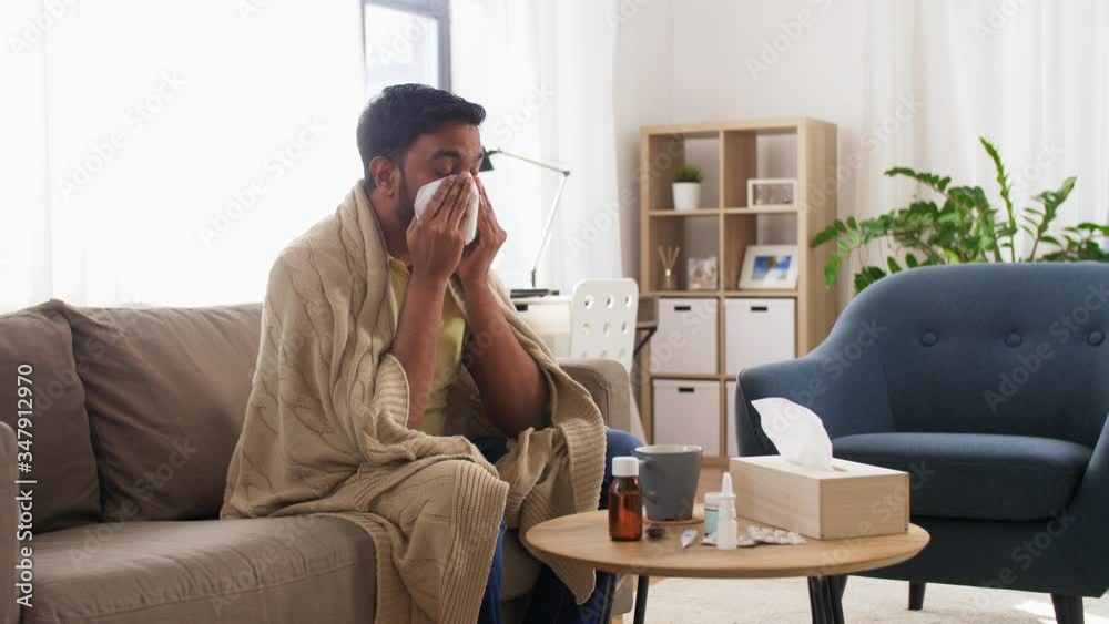 healthcare, cold, hygiene and people concept - sick indian man in blanket sneezing and blowing nose with paper tissue at home