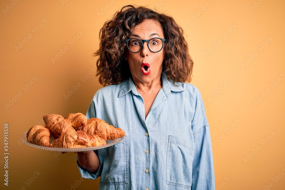 Middle age beautiful woman holding plate with croissants over isolated yellow background scared in shock with a surprise face, afraid and excited with fear expression Middle age beautiful woman holding plate with croissants over isolated yellow background scared in shock with a surprise face, afraid and excited with fear expression