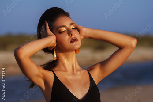 Portrait of a tanned model by the sea with a blurred background. beautiful evening light. makeup with gold