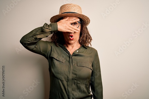 Middle age brunette woman wearing glasses and hat standing over isolated white background peeking in shock covering face and eyes with hand, looking through fingers with embarrassed expression.