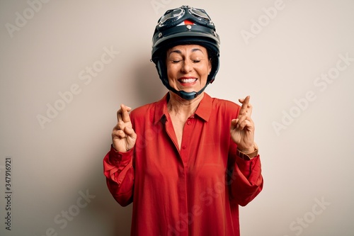 Middle age motorcyclist woman wearing motorcycle helmet over isolated white background gesturing finger crossed smiling with hope and eyes closed. Luck and superstitious concept.