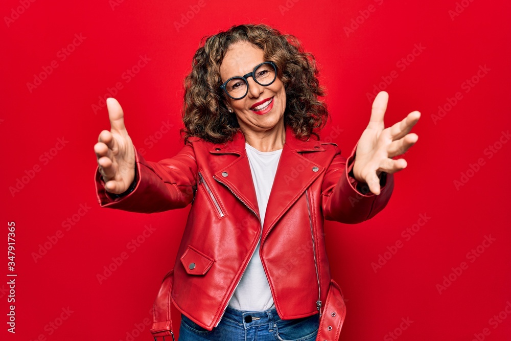 Middle age beautiful woman wearing casual red jacket and glasses over isolated background looking at the camera smiling with open arms for hug. Cheerful expression embracing happiness.