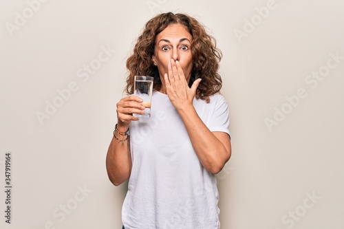 Middle age beautiful woman drinking glass of water to refreshment over white background covering mouth with hand, shocked and afraid for mistake. Surprised expression © Krakenimages.com