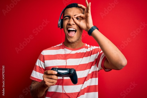 Handsome african american gamer man playing video game using jostick and headphones with happy face smiling doing ok sign with hand on eye looking through fingers photo