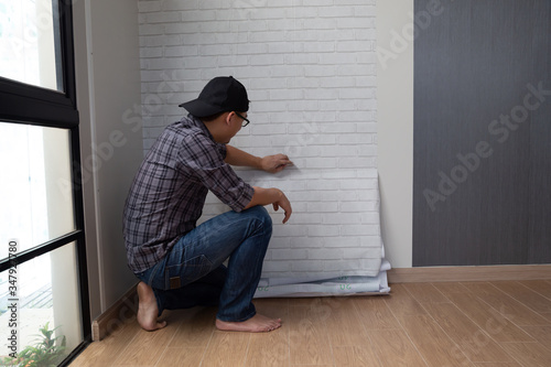 Young Asian man glueing wallpapers on the wall at home. renovation room in house by self diy concept