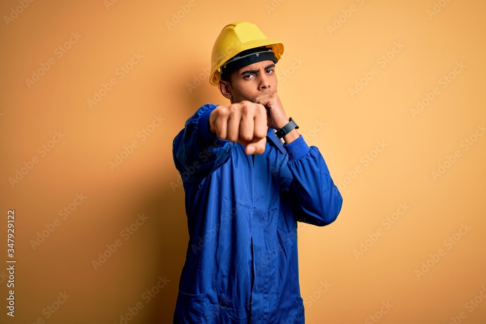 Young handsome african american worker man wearing blue uniform and security helmet Punching fist to fight, aggressive and angry attack, threat and violence
