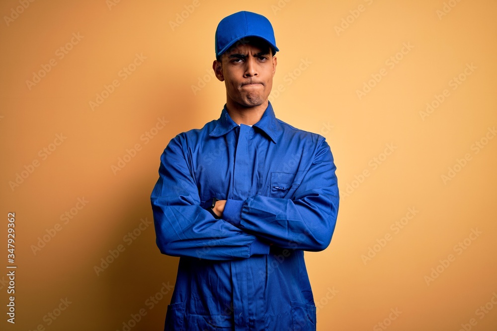 Young african american mechanic man wearing blue uniform and cap over yellow background skeptic and nervous, disapproving expression on face with crossed arms. Negative person.