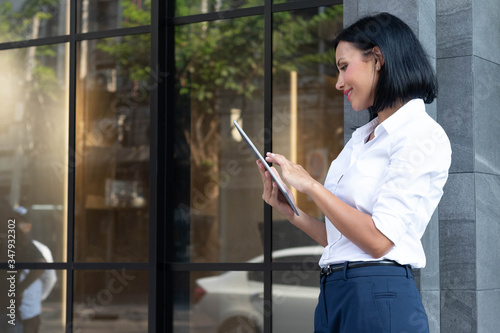 Half figure portrait of smart succdessful businesswoman smiling, using tablet computer infront of office building in downtown urban area and ready to work.