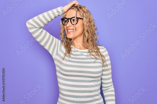 Beautiful blonde woman wearing casual striped t-shirt and glasses over purple background smiling confident touching hair with hand up gesture, posing attractive and fashionable