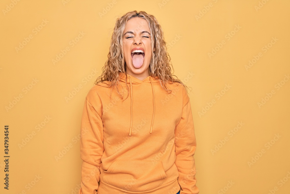 Young beautiful blonde sporty woman wearing casual sweatshirt over yellow background sticking tongue out happy with funny expression. Emotion concept. Young beautiful blonde sporty woman wearing casual sweatshirt over yellow background sticking tongue out happy with funny expression. Emotion concept.