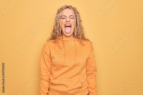 Young beautiful blonde sporty woman wearing casual sweatshirt over yellow background sticking tongue out happy with funny expression. Emotion concept. © Krakenimages.com