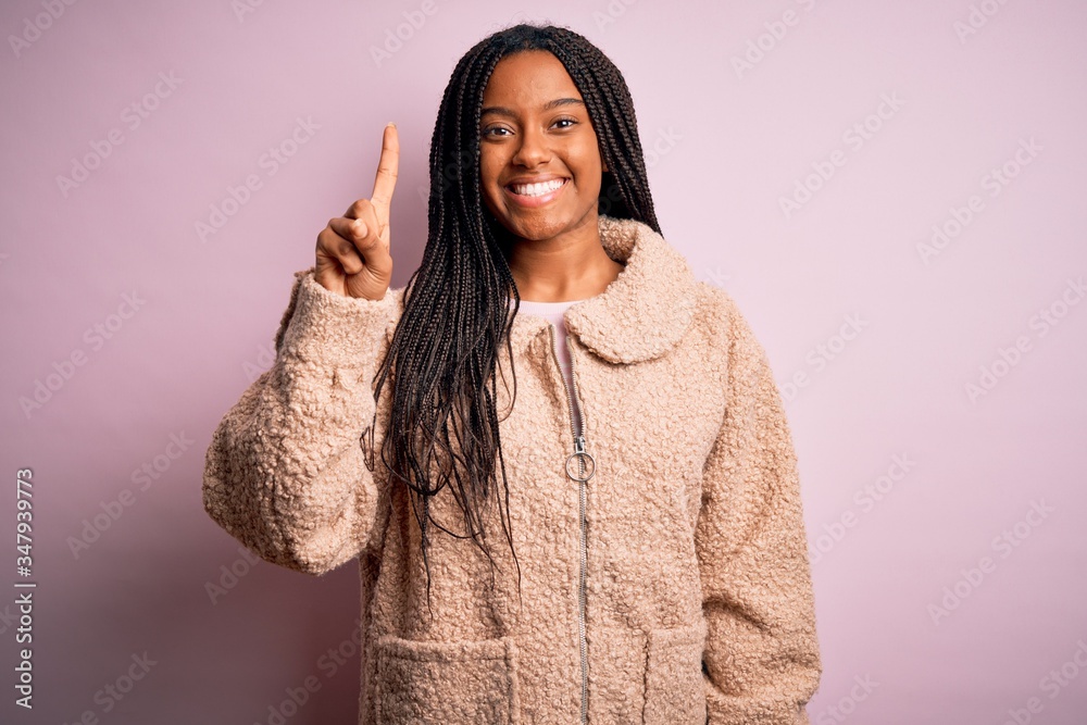 Young african american woman wearing fashion winter coat over pink isolated background showing and pointing up with finger number one while smiling confident and happy. Young african american woman wearing fashion winter coat over pink isolated background showing and pointing up with finger number one while smiling confident and happy.