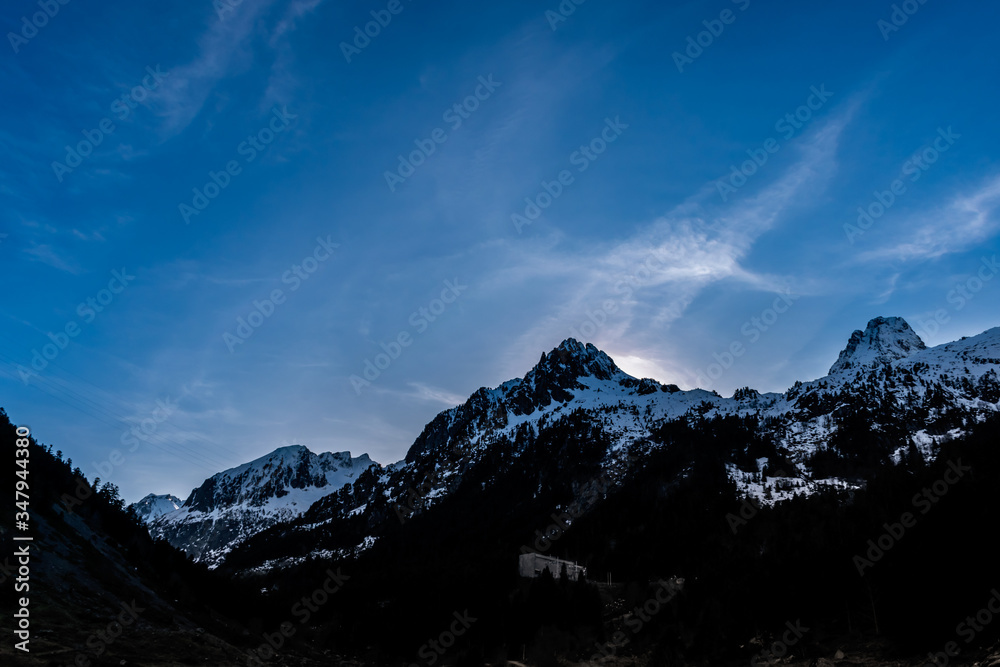 A picturesque landscape view of the high snow capped Pyrenees mountains on a winter evening with the sun behind the peak and a clear blue sky (Arrens-Marsous, Hautes-Pyrenees, France)