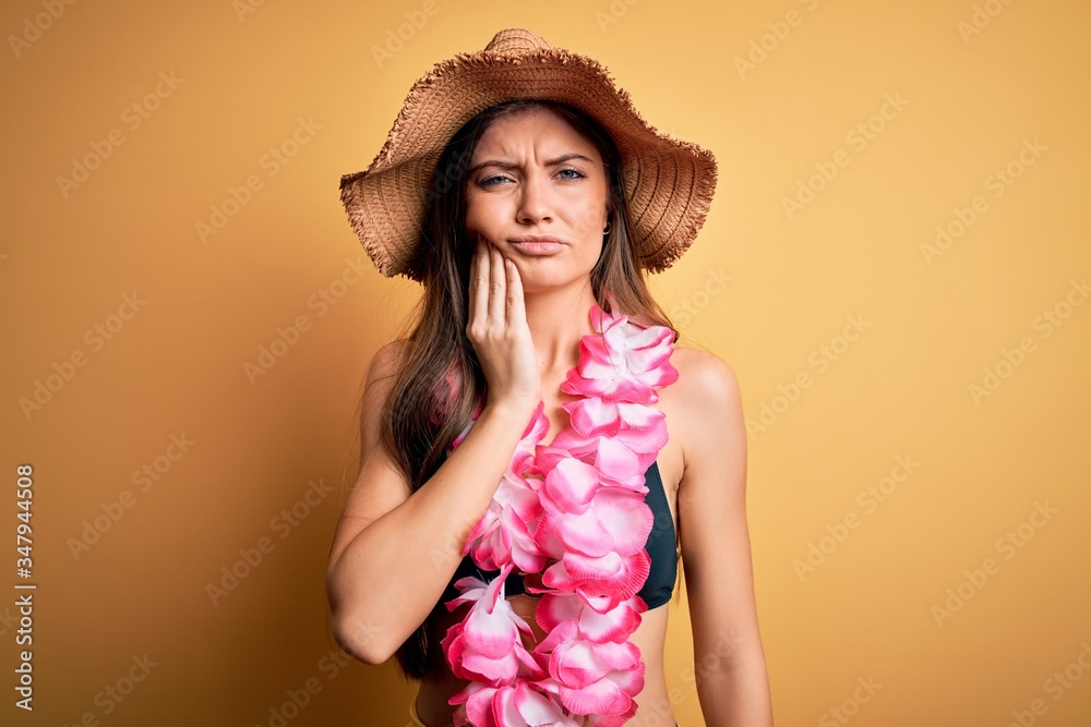 Young beautiful woman with blue eyes on vacation wearing bikini and hawaiian lei touching mouth with hand with painful expression because of toothache or dental illness on teeth. Dentist Young beautiful woman with blue eyes on vacation wearing bikini and hawaiian lei touching mouth with hand with painful expression because of toothache or dental illness on teeth. Dentist