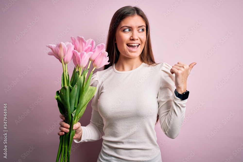 Young beautiful romantic woman with blue eyes holding beauty bouquet of pink tulips pointing and showing with thumb up to the side with happy face smiling Young beautiful romantic woman with blue eyes holding beauty bouquet of pink tulips pointing and showing with thumb up to the side with happy face smiling