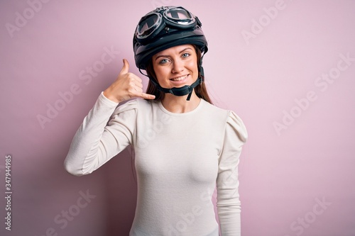 Young beautiful motorcyclist woman with blue eyes wearing moto helmet over pink background smiling doing phone gesture with hand and fingers like talking on the telephone. Communicating concepts.