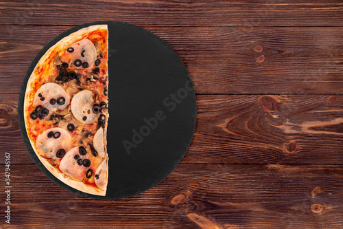 Half a pizza with ham, mozzarella, mushrooms and olives on a slate platter which is on wooden background, top view and copy space