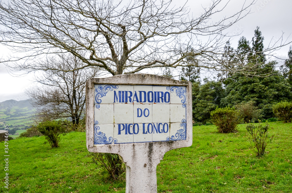 Blue sign Miradoiro do Pico Longo, Viewpoint Pico Longo in English, on typical Portuguese tiles. Grass and trees without leaves in the background. Horizontal photo.