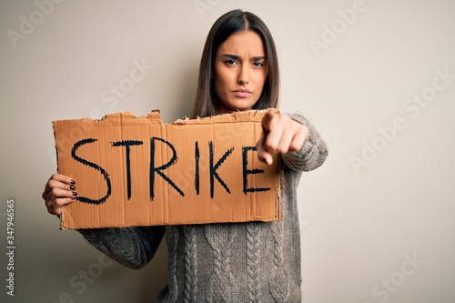 Young beautiful brunette activist woman protesting holding poster with strike message pointing with finger to the camera and to you, hand sign, positive and confident gesture from the front