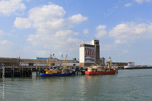 the fishing port of breskens, zeeland, the netherlands in summer with two fishing boats and a grain silo with an artwork painting of breads and fish in the background