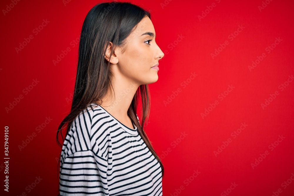 Young beautiful brunette woman wearing casual striped t-shirt over red background looking to side, relax profile pose with natural face with confident smile. Young beautiful brunette woman wearing casual striped t-shirt over red background looking to side, relax profile pose with natural face with confident smile.