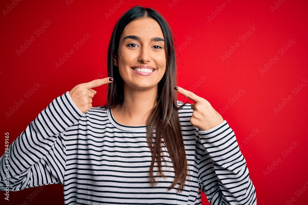 Young beautiful brunette woman wearing casual striped t-shirt over red background smiling cheerful showing and pointing with fingers teeth and mouth. Dental health concept. Young beautiful brunette woman wearing casual striped t-shirt over red background smiling cheerful showing and pointing with fingers teeth and mouth. Dental health concept.