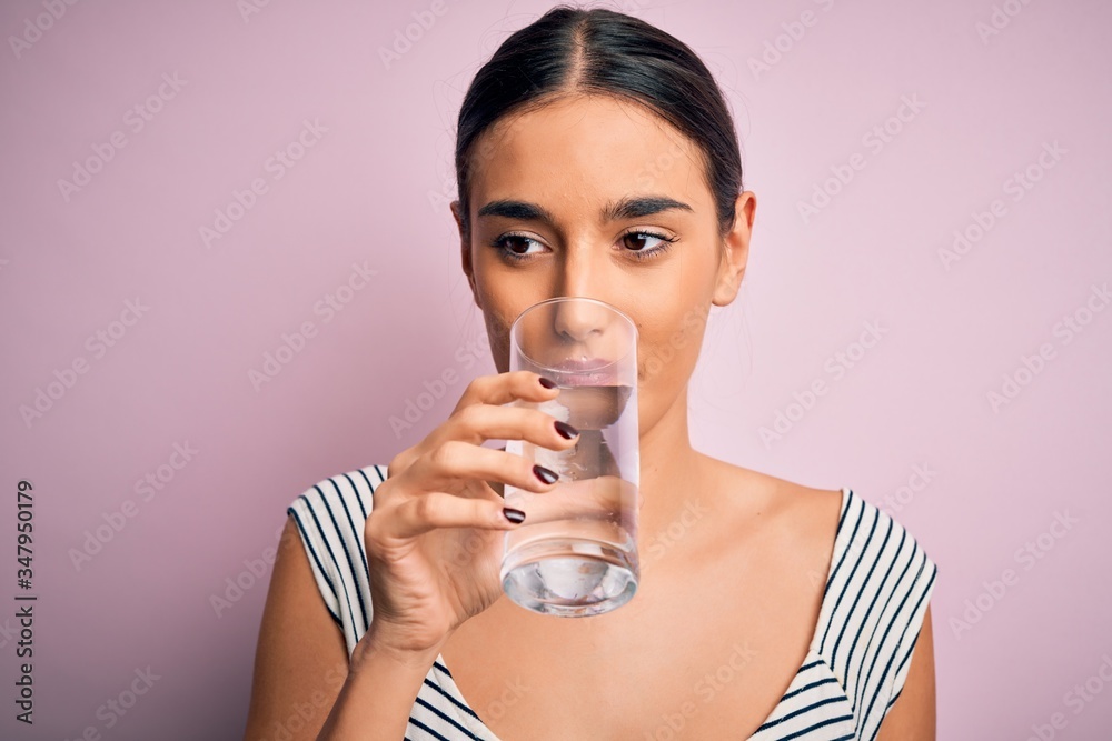Young beautiful brunette woman drinking glass of healthy water to refreshment standing over isolated pink background Young beautiful brunette woman drinking glass of healthy water to refreshment standing over isolated pink background