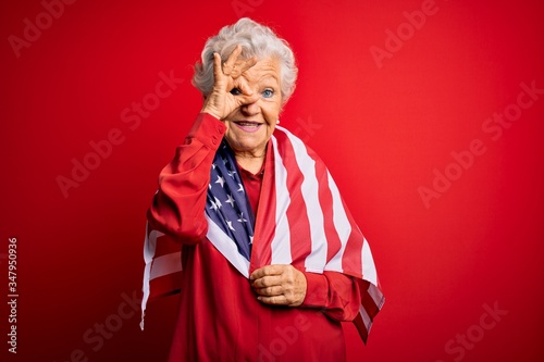 Senior beautiful grey-haired patriotic woman wearing united states flag over red background with happy face smiling doing ok sign with hand on eye looking through fingers