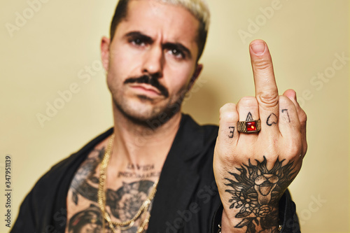 Young aggressive provocative male in black coat over naked tattooed torso showing middle finger gesture while standing against beige background