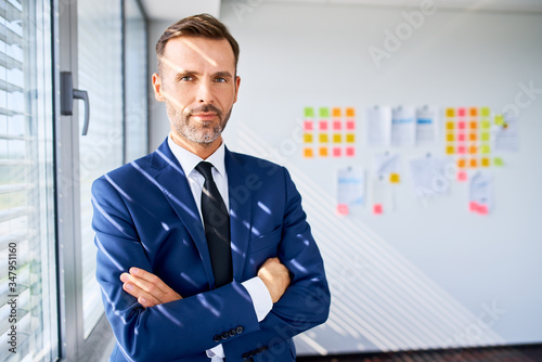 Portrait of confident adult businessman standing at office with crossed arms, looking at camera