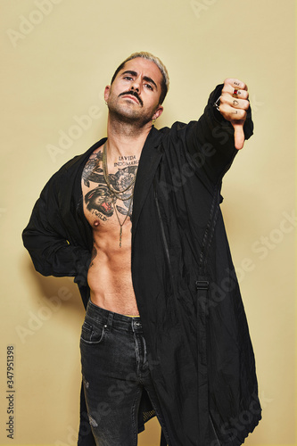 Young provocative male in black coat over naked tattooed torso showing thumbs down gesture in disapproval while standing against beige background photo