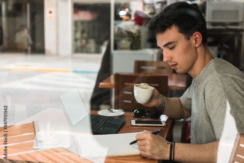 Side view of young man with cup of coffee writing in notepad while sitting at table behind glass and studying in cafeteria Side view of young man with cup of coffee writing in notepad while sitting at table behind glass and studying in cafeteria