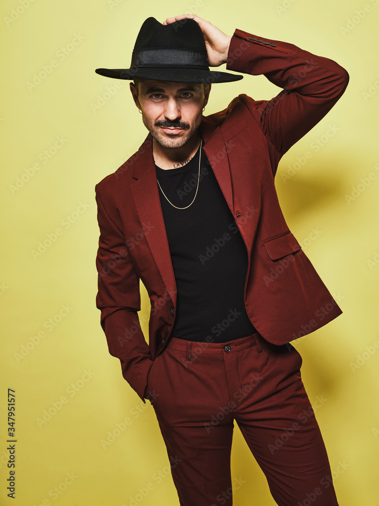 Handsome bearded well dressed male in trendy vinous suit and hat looking at camera against yellow background Handsome bearded well dressed male in trendy vinous suit and hat looking at camera against yellow background