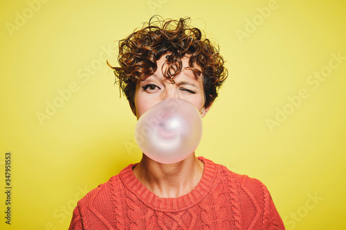 Young curly haired female in orange sweater blowing bubble gum and blinking looking to the camera while standing against yellow background