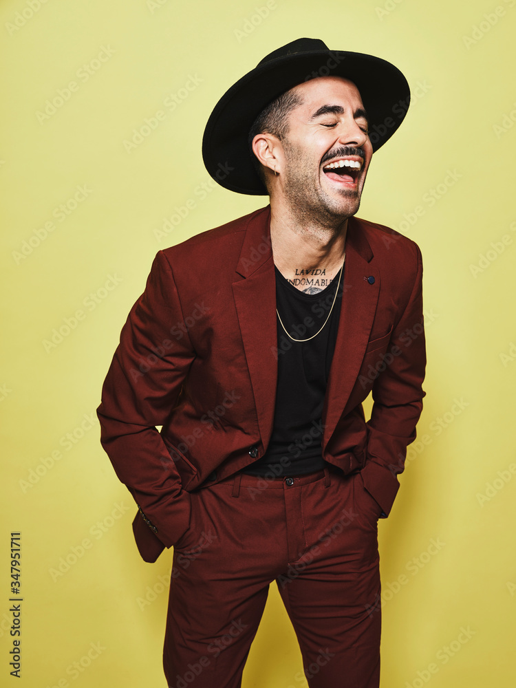 Laughing adult ethnic male in trendy vinous suit and hat with closed eyes standing against yellow background Laughing adult ethnic male in trendy vinous suit and hat with closed eyes standing against yellow background