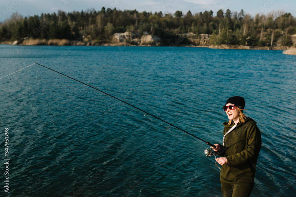 Woman catching a fish, pulling rod while fishing on lake or pond at the weekend. Fisherman with rod, spinning reel on the river bank. Sunrise. Fishing for pike, perch, carp. Background wild nature. Woman catching a fish, pulling rod while fishing on lake or pond at the weekend. Fisherman with rod, spinning reel on the river bank. Sunrise. Fishing for pike, perch, carp. Background wild nature.