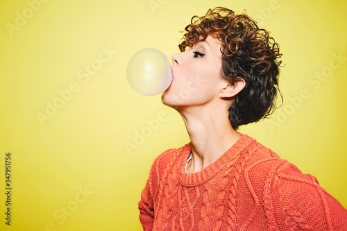Side view of young pretty curly haired female in orange sweater blowing bubble gum while standing looking away against yellow background