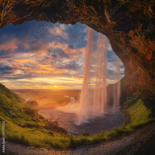 Majestic waterfall located on entrance of grassy stone cave against cloudy sundown sky on seashore in Iceland