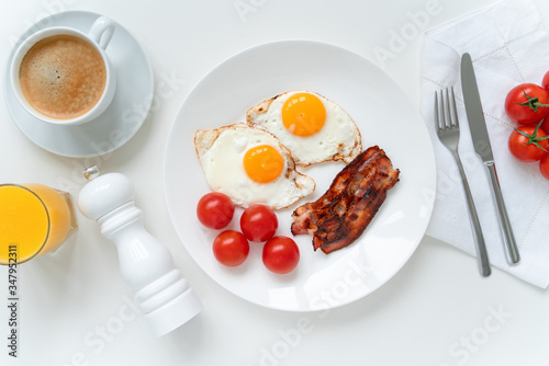 American breakfast with two eggs, bacon, cherry tomatoes, coffee and orange juice on the white table. Top view.