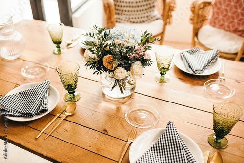 From above bouquet of miscellaneous flowers and green plant twigs in vase with water on a wooden table set for a meal