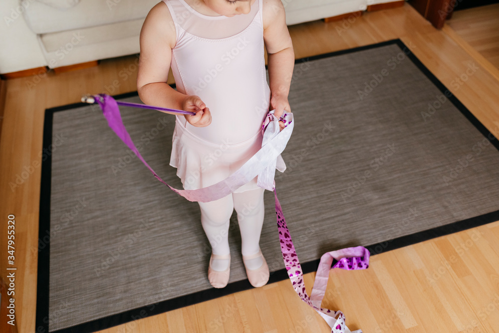 From above focused cropped cute little girl in leotard and tights spinning ribbon during rhythmic gymnastic practice training in cozy living room at home From above focused cropped cute little girl in leotard and tights spinning ribbon during rhythmic gymnastic practice training in cozy living room at home