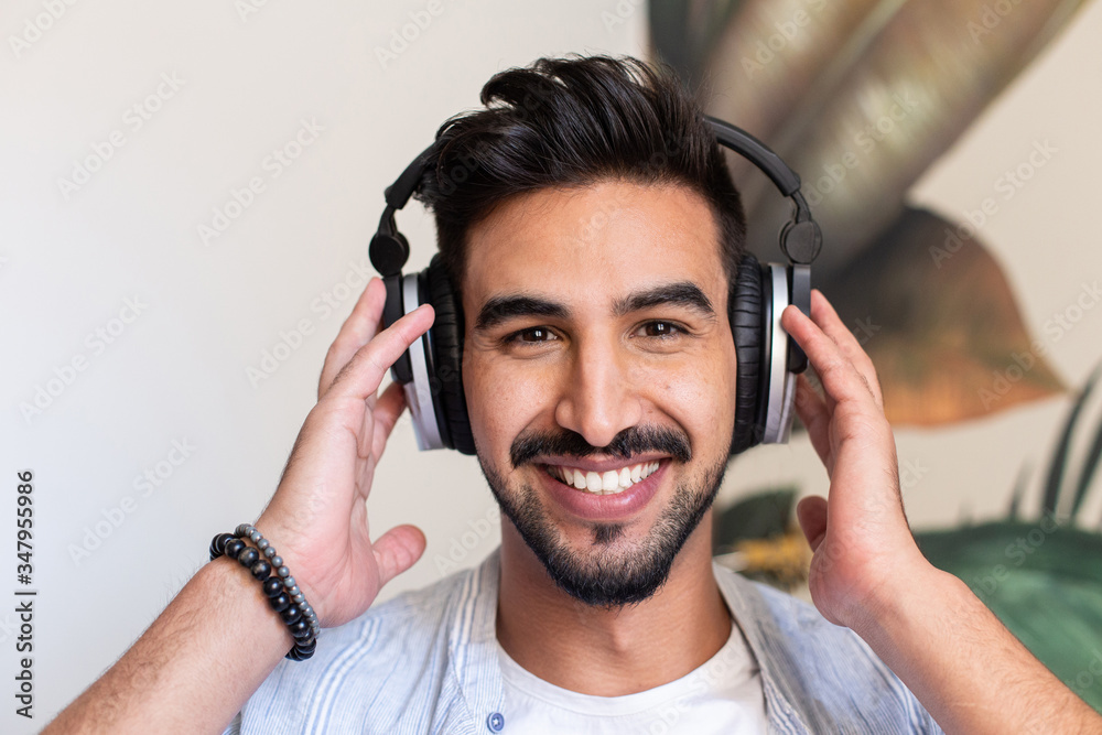 Happy ethnic guy in headphones smiling and looking at camera while listening to music at home Happy ethnic guy in headphones smiling and looking at camera while listening to music at home
