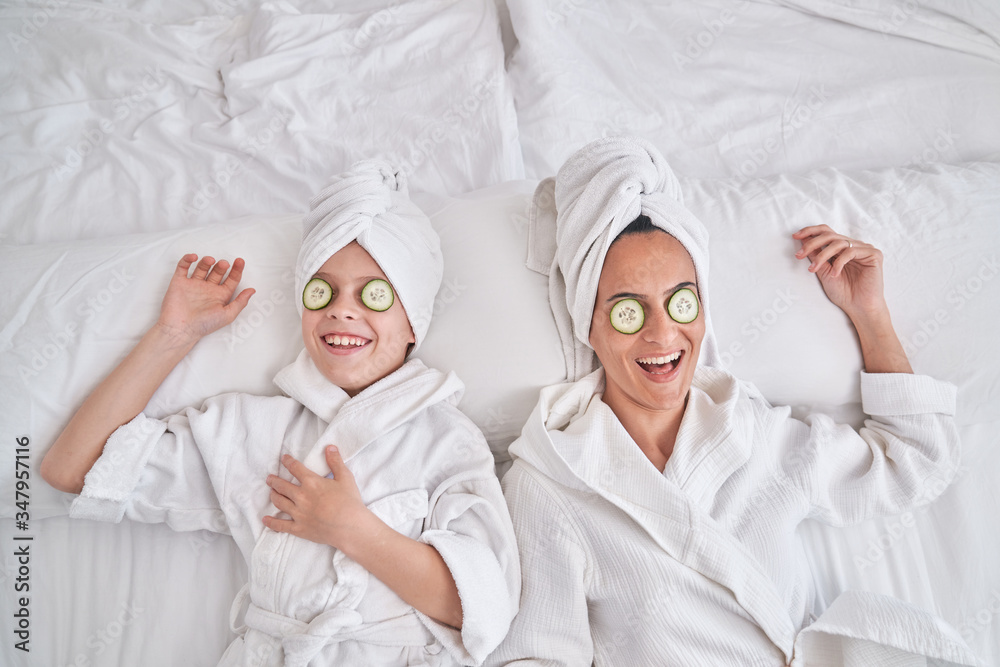 From above of happy young female and kid wearing white bathrobes and towel turbans lying on bed with cucumber slices on eyes and laughing while having fun and enjoying beauty procedure at home From above of happy young female and kid wearing white bathrobes and towel turbans lying on bed with cucumber slices on eyes and laughing while having fun and enjoying beauty procedure at home