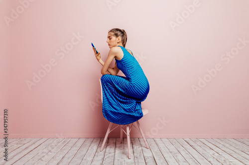 Side view of young woman crouched on top of a chair while using mobile phone on pink background