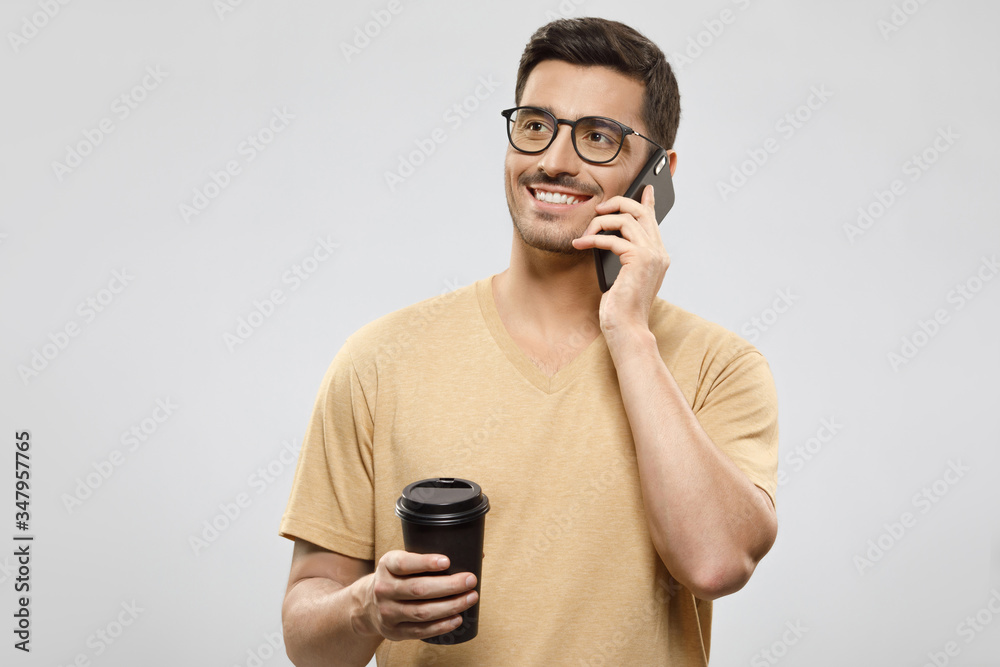 Young man in t-shirt and glasses, talking on the phone with positive smile, holding takeaway coffee cup, isolated on gray studio background Young man in t-shirt and glasses, talking on the phone with positive smile, holding takeaway coffee cup, isolated on gray studio background